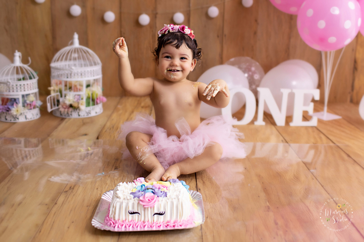 Ensaio Smash the cake da Laura de 1 ano em studio localizado na Vila Mariana - São Paulo, SP. Na foto a bebê está sentada com seu cenário rosa com bexigas e flores, dando risada e brincando com seu lindo bolo de unicórnio