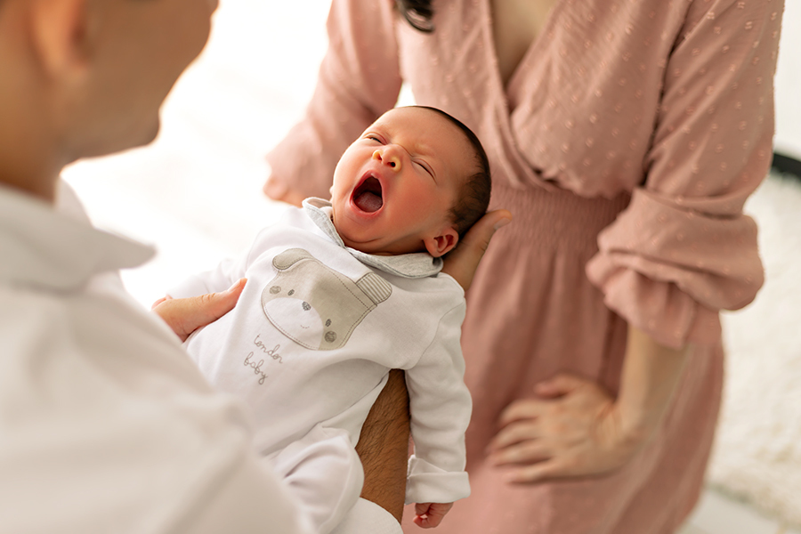 Foto de recém-nascido sendo segurada pelo pai. Mãe ao fundo, bebê bocejando em destaque.