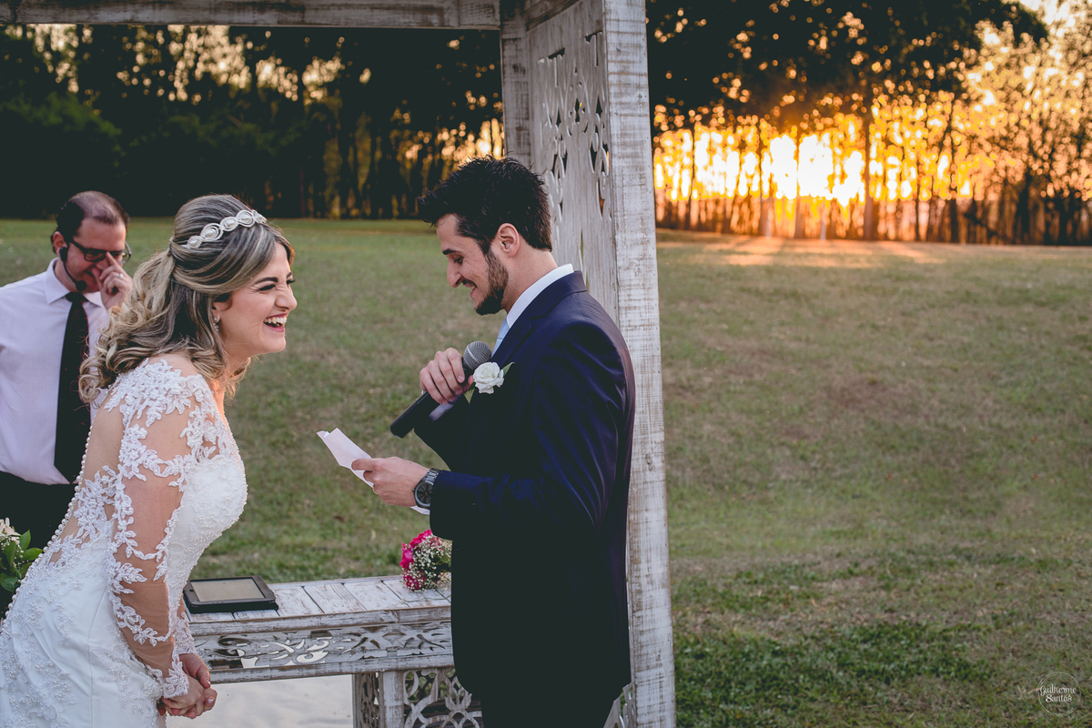 Fotografia de casamento pelo fotógrafo Guilherme Santos, noiva sorrindo durante os votos do noivo na cerimônia de casamento em Pederneiras. Casamento ao ar livre no pôr do sol.