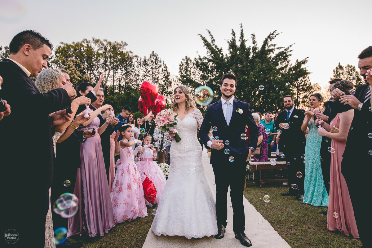Fotografia de casamento pelo fotógrafo Guilherme Santos, casal de noivos sorrindo durante a saída do casamento ao ar livre no fim de tarde em Pederneiras. Bola de sabão na saída nos noivos.