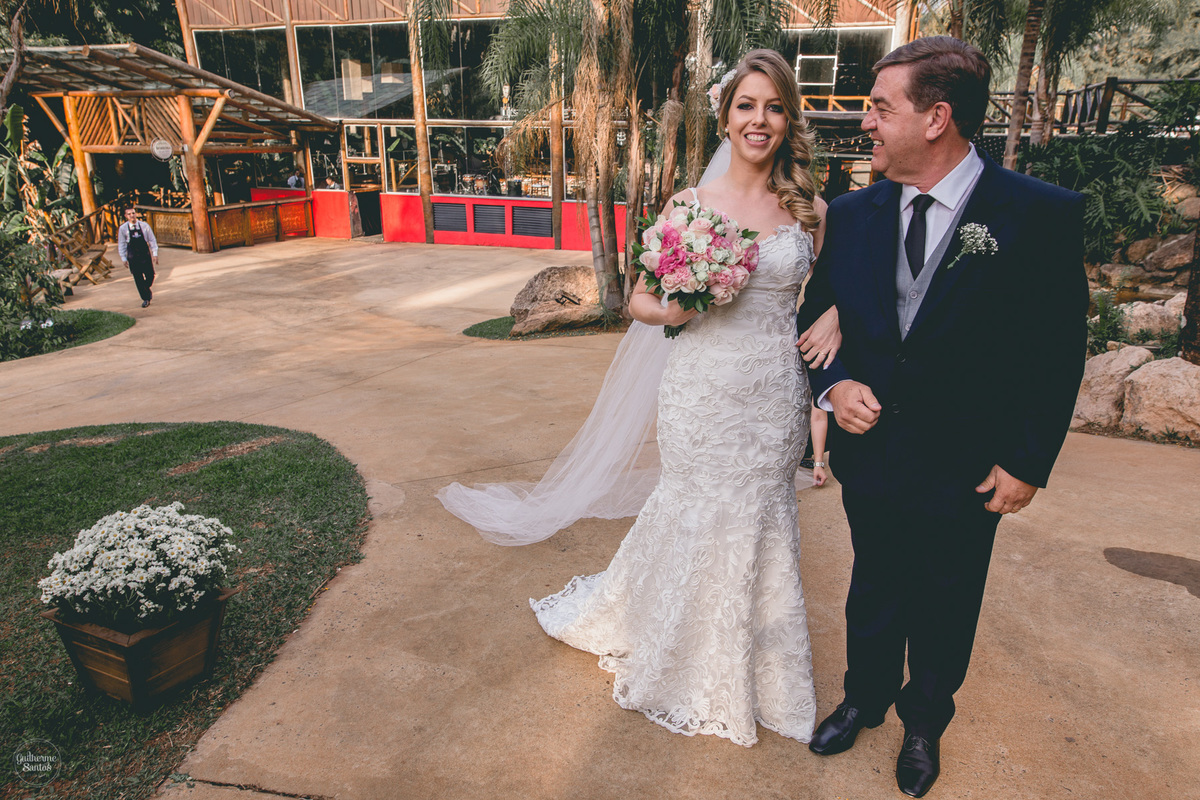 Fotografia de casamento feita no final de tarde pelo fotógrafo Guilherme Santos, noiva com seu pai. Vestido da noiva lindo, lugar encantador. Noiva está sorrindo e segurando o buquê.