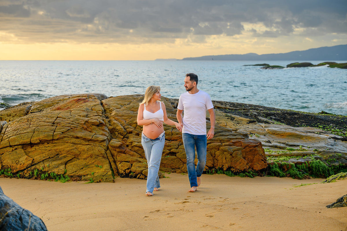 Ensaio fotográfico de gestante ao amanhecer em Balneário Camboriú com casal caminhando pela areia da praia.