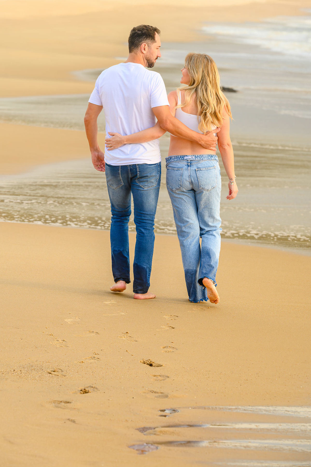 Ensaio fotográfico de gestante ao amanhecer em Balneário Camboriú com casal caminhando pela areia da praia.