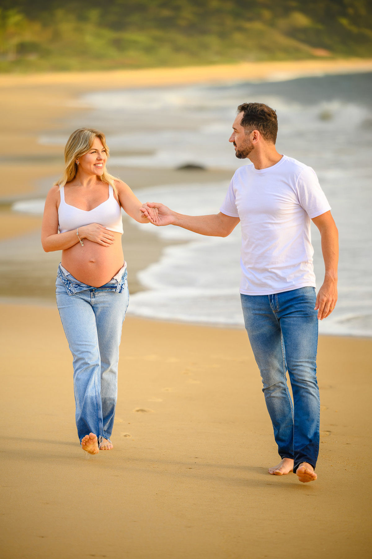 Ensaio fotográfico de gestante ao amanhecer em Balneário Camboriú com casal caminhando pela areia da praia.
