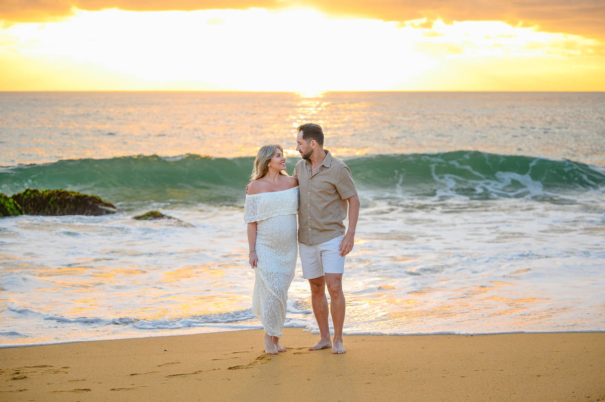Ensaio fotográfico de gestante ao amanhecer em Balneário Camboriú com casal caminhando pela areia da praia.
