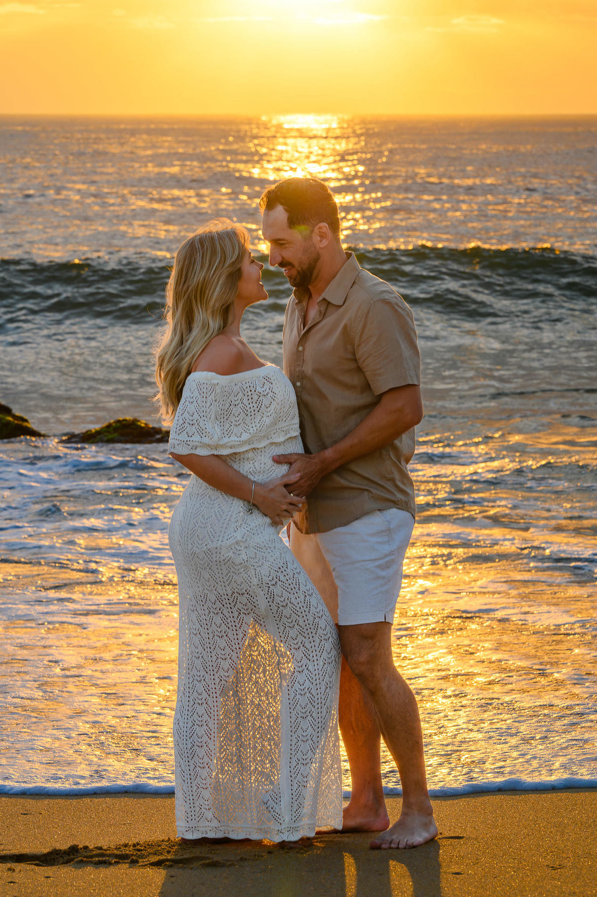 Gestante e parceiro em ensaio na Praia do Estaleirinho ao amanhecer, com mar calmo e luz natural da manhã.