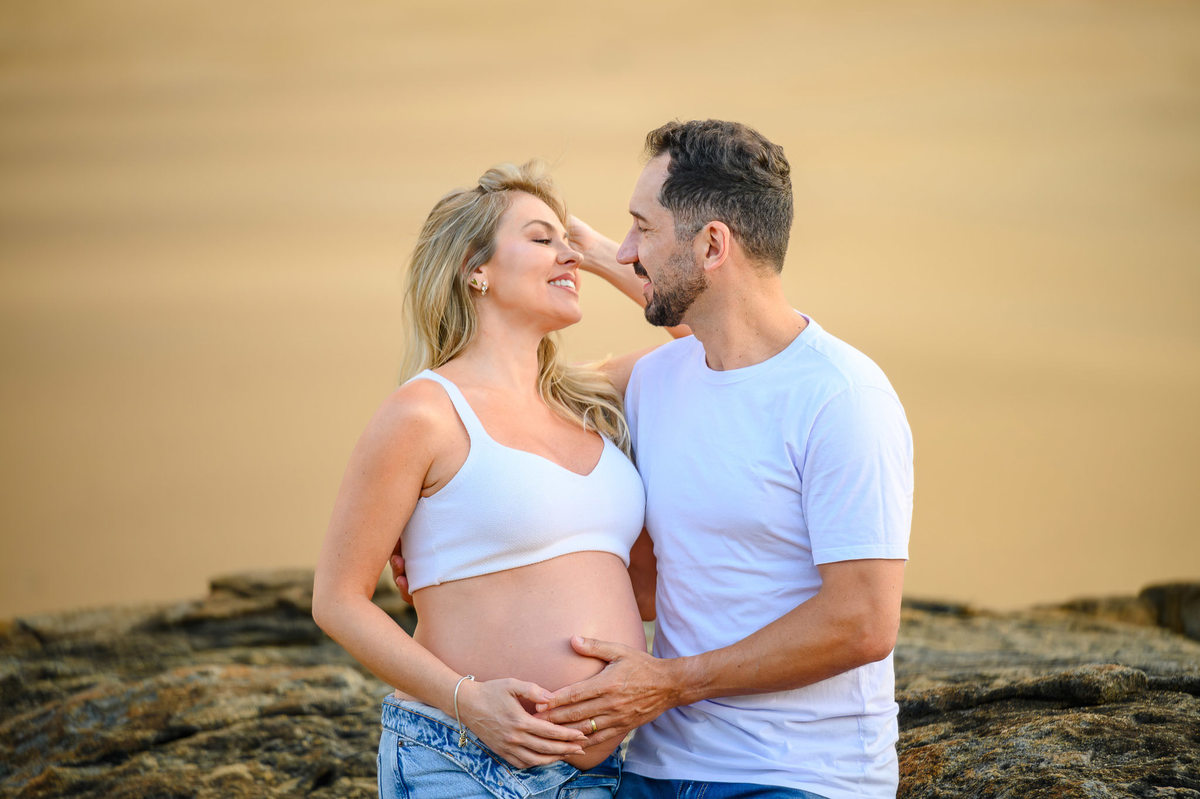 Gestante e parceiro em ensaio na Praia do Estaleirinho ao amanhecer, com mar calmo e luz natural da manhã.