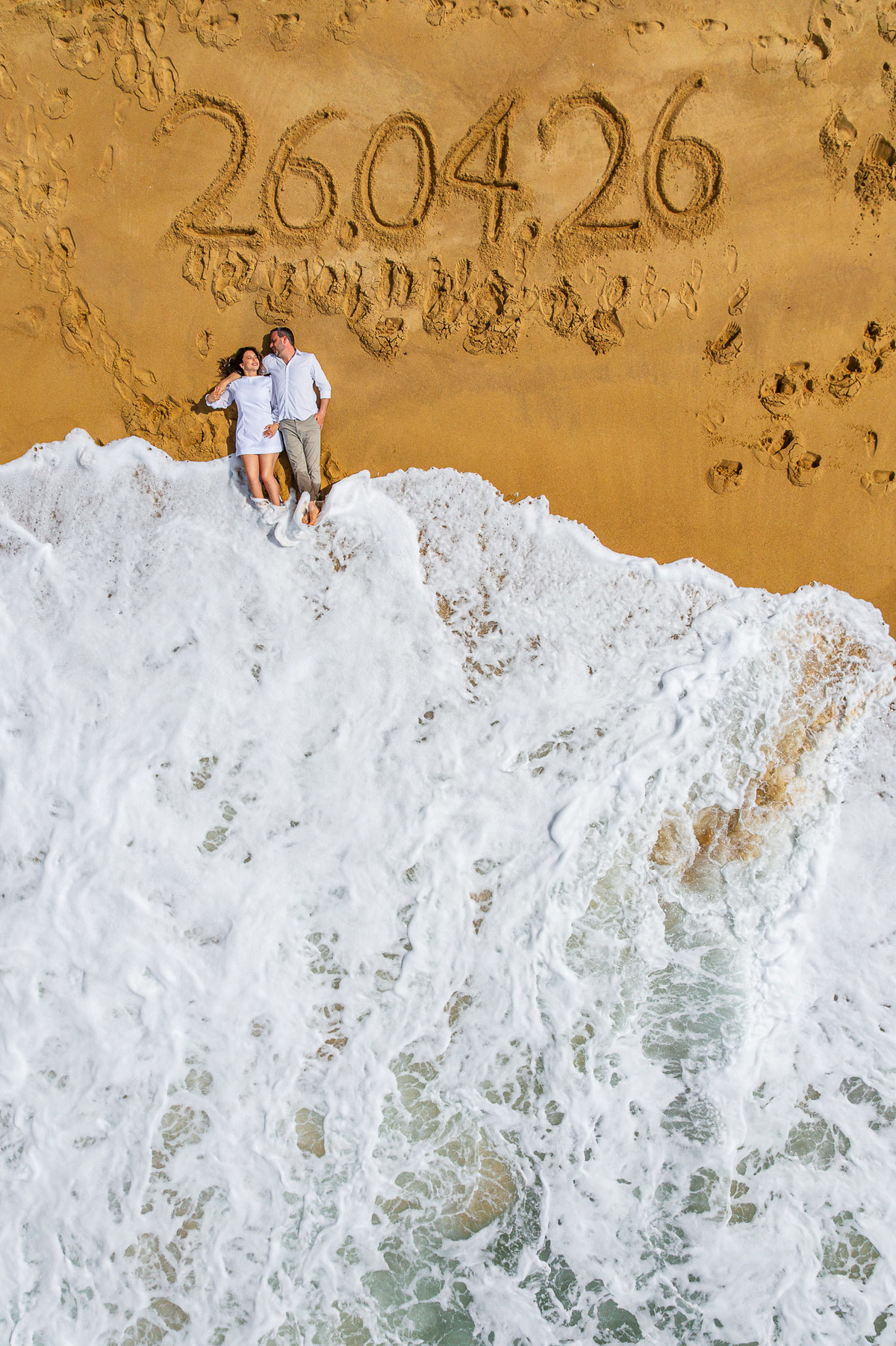 Foto aérea com drone de casal no pré-wedding na praia com data escrita na areia