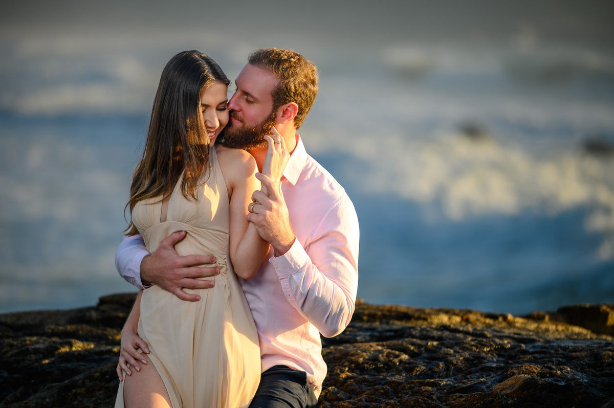 Casal em momento íntimo na praia com cenário de rochas e mar ao amanhecer