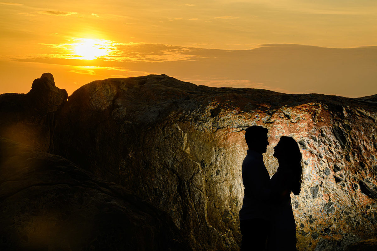 Silhueta de casal em frente a pedras com luz dramática ao amanhecer na Praia do Estaleirinho – SC