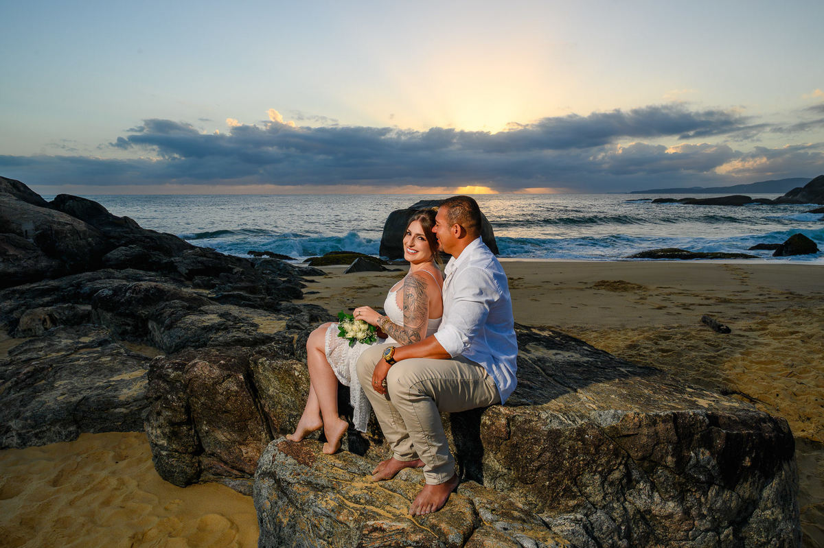 Casal sentado nas rochas da praia ao amanhecer com o mar ao fundo, ensaio romântico em Balneário Camboriú