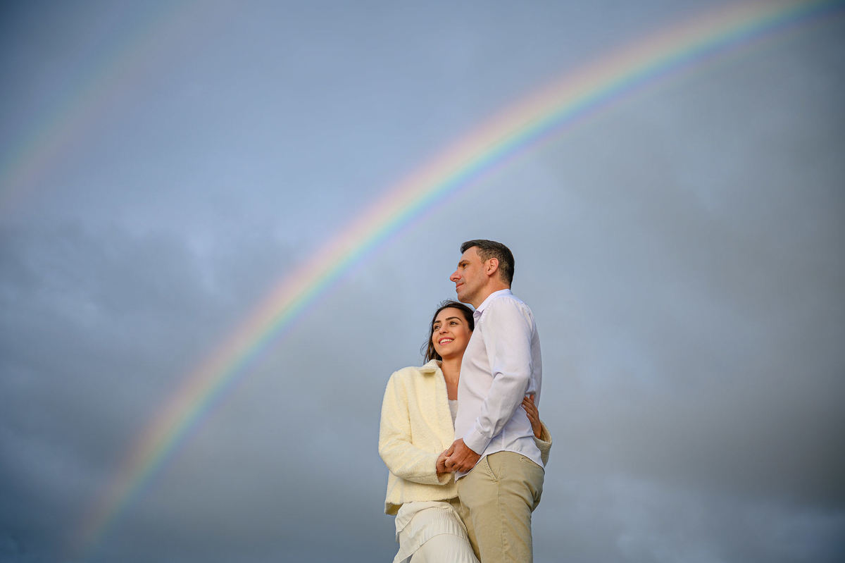 Casal em pé sobre as pedras com arco-íris ao fundo durante ensaio pré-wedding em Balneário Camboriú SC