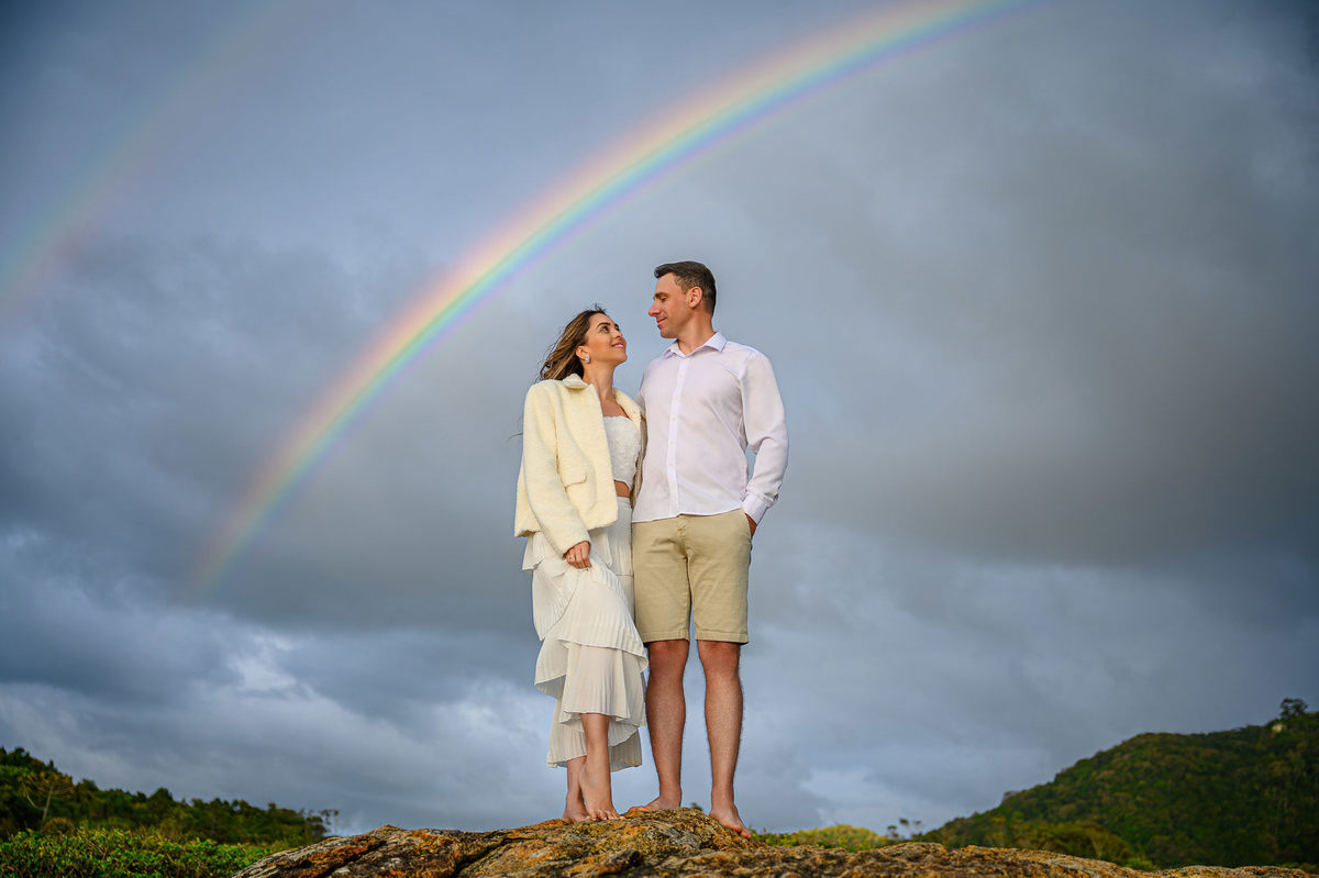 Casal em pé sobre as pedras com arco-íris ao fundo durante ensaio pré-wedding em Balneário Camboriú SC