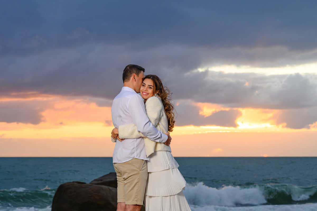 Ensaio pré-wedding ao amanhecer na Praia do Estaleirinho em Balneário Camboriú SC com casal abraçado diante do mar e das pedras