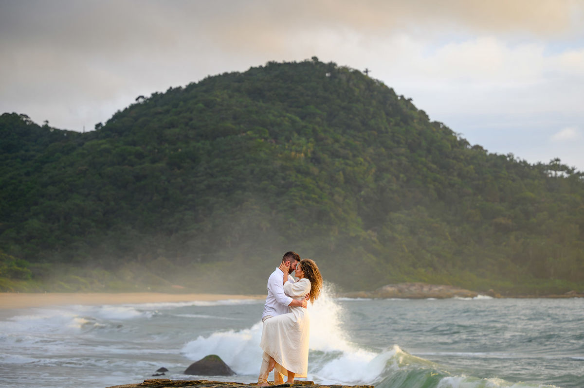 Casal abraçado próximo às pedras com ondas do mar ao fundo durante ensaio pré-wedding ao amanhecer na Praia do Estaleirinho.