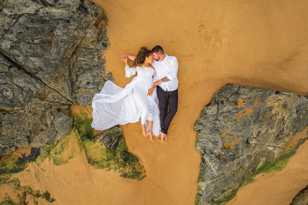 Casal deitado na areia entre pedras visto de cima durante ensaio pré-wedding ao amanhecer na Praia do Estaleirinho, Santa Catarina.