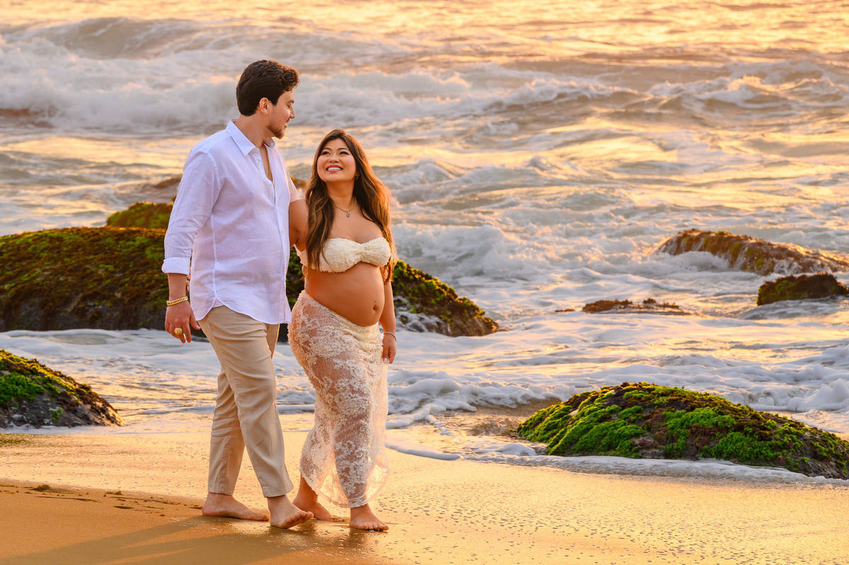 Ensaio fotográfico de gestante ao amanhecer em Balneário Camboriú com casal caminhando pela areia da praia.