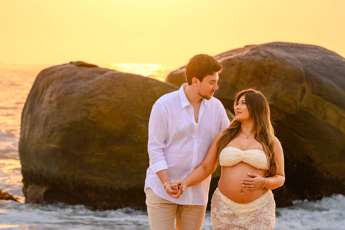 Ensaio fotográfico de gestante ao amanhecer em Balneário Camboriú com casal caminhando pela areia da praia.