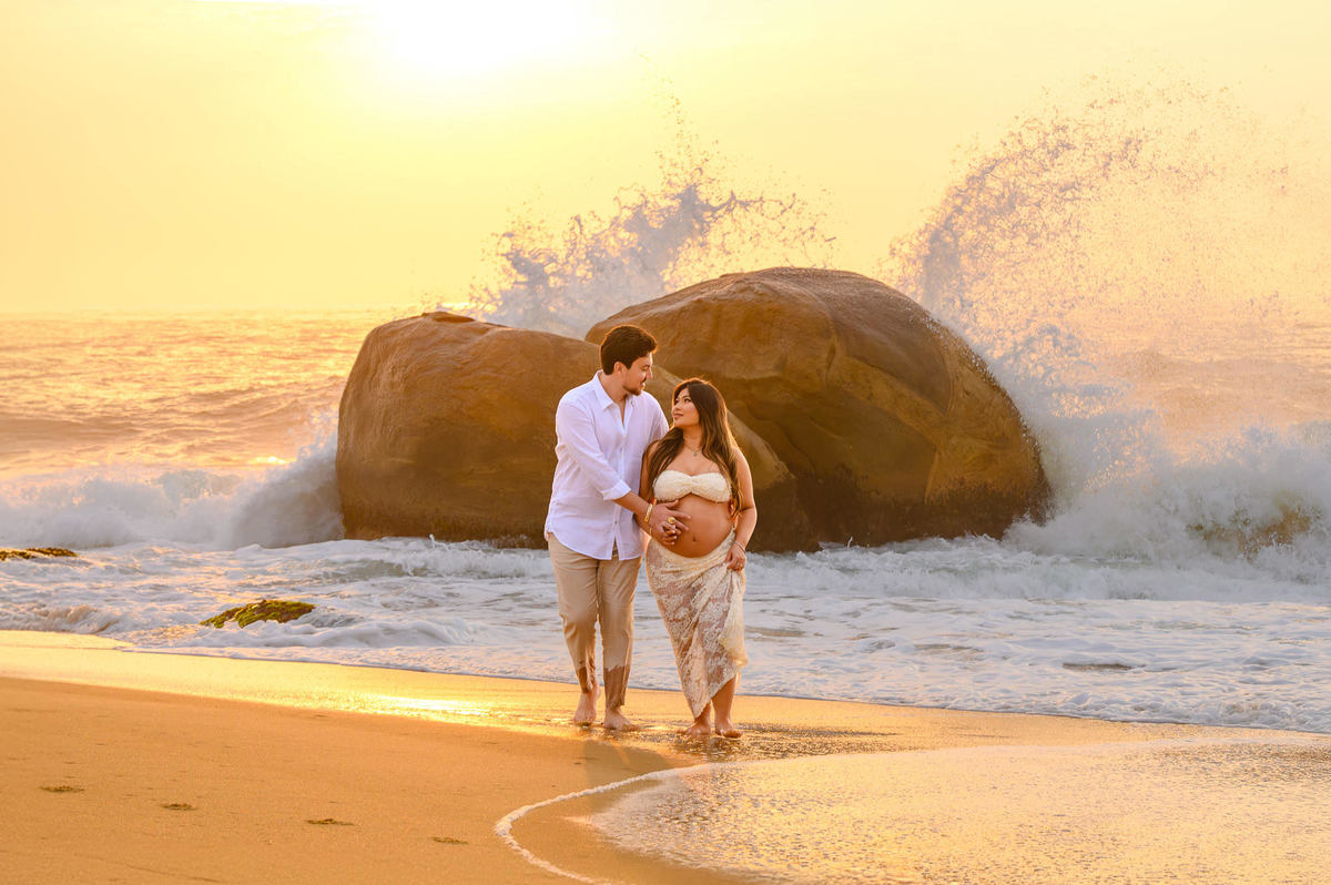 Ensaio fotográfico de gestante ao amanhecer em Balneário Camboriú com casal caminhando pela areia da praia.