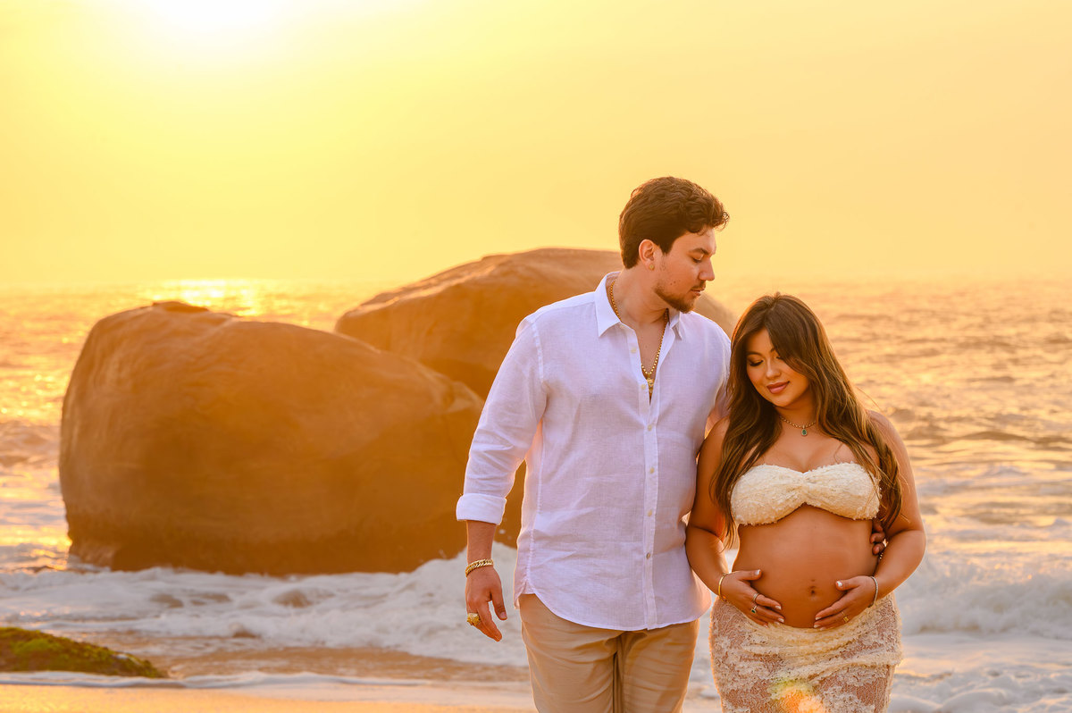 Ensaio fotográfico de gestante ao amanhecer em Balneário Camboriú com casal caminhando pela areia da praia.