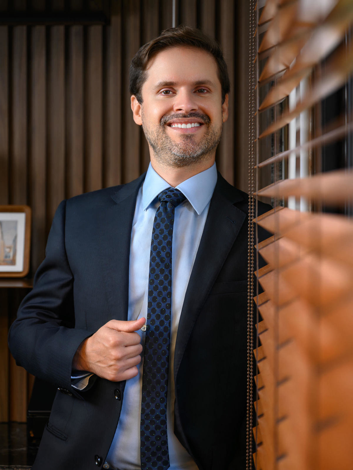 Retrato profissional masculino em ambiente corporativo com persianas e iluminação suave, transmitindo seriedade e autoridade.