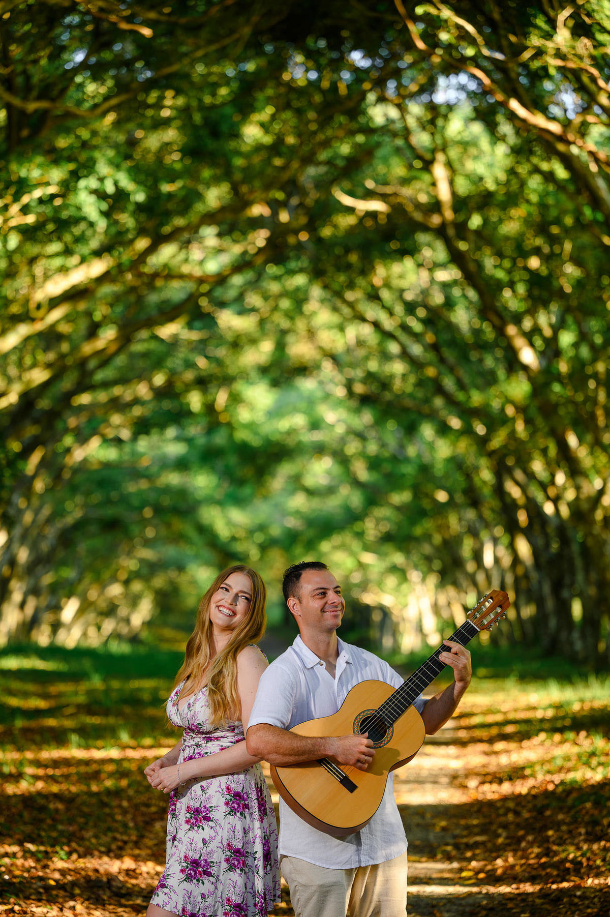 Casal em trilha no bosque, ele tocando violão enquanto ela sorri ao lado, luz natural filtrada pelas árvores