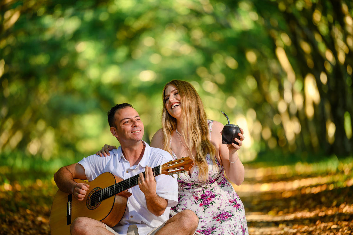 Casal sentado no bosque, momento descontraído com violão, rodeado por árvores formando um túnel natural verde