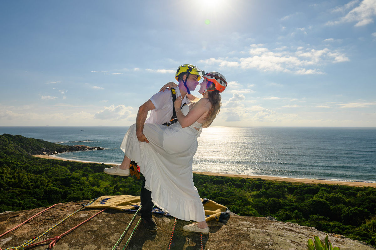 Casal se abraçando em um mirante natural com vista para o mar, usando equipamentos de rappel, ensaio pré-wedding de aventura em Balneário Camboriú, SC