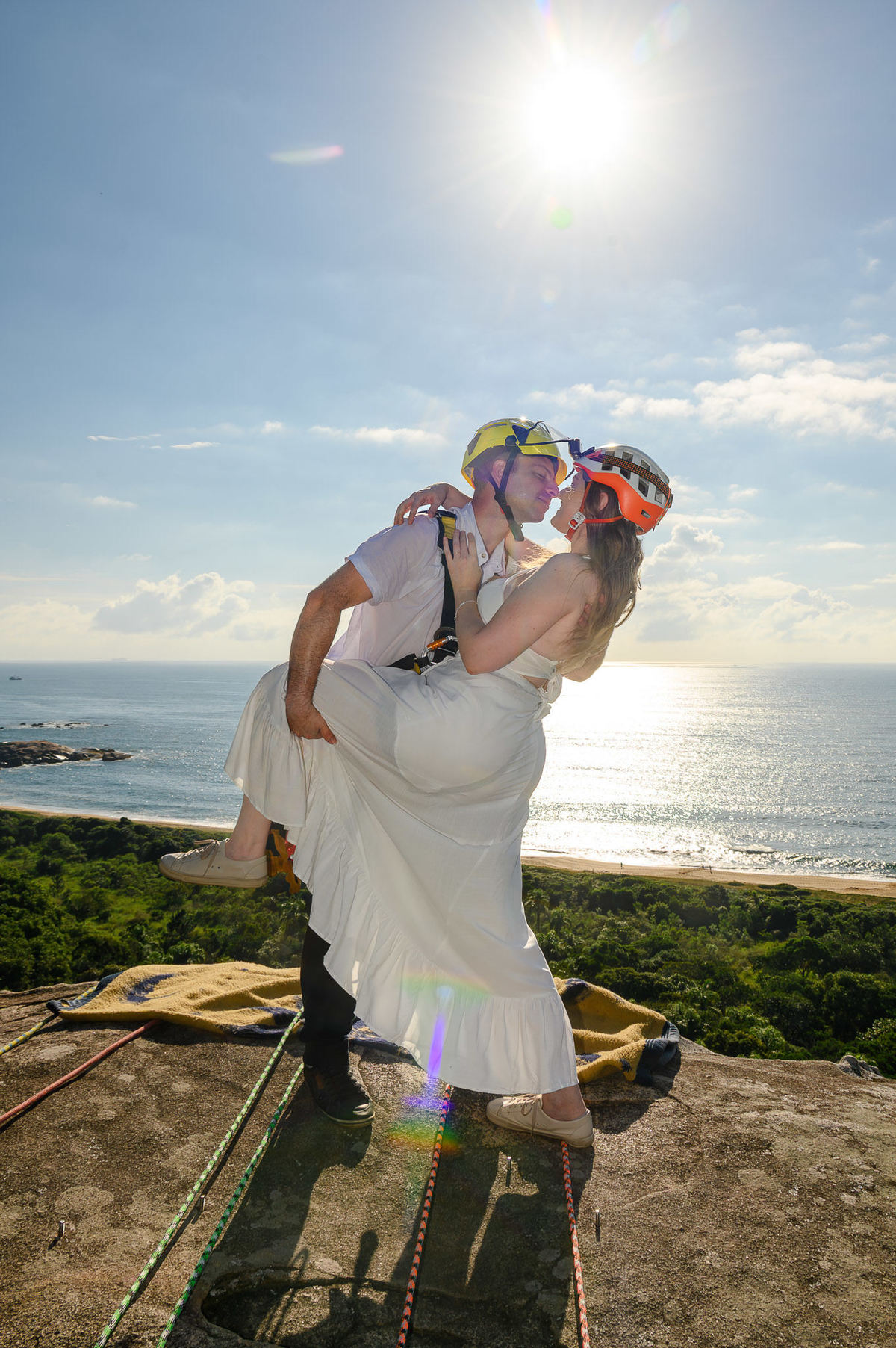 Casal com equipamentos de rappel abraçados no topo da rocha, com o oceano ao fundo e luz natural do início do dia