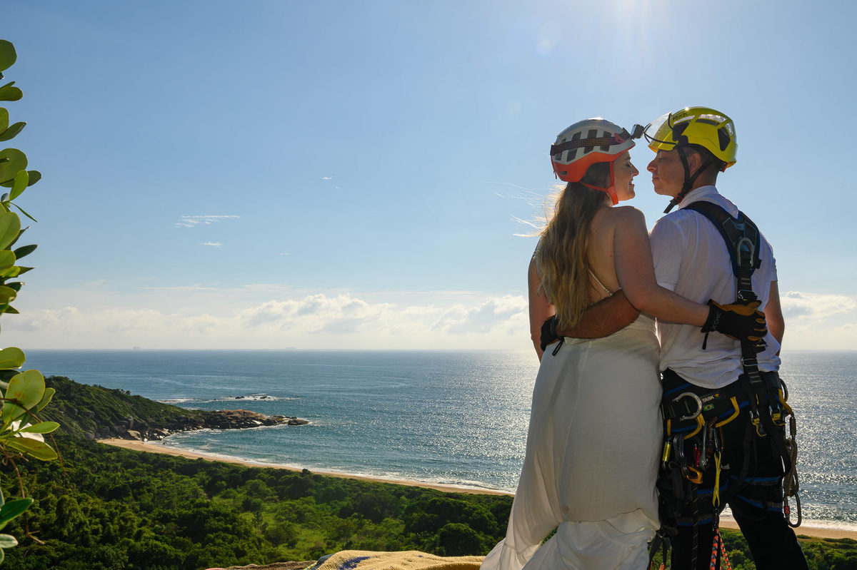 Casal com equipamentos de rappel abraçados no topo da rocha, com o oceano ao fundo e luz natural do início do dia