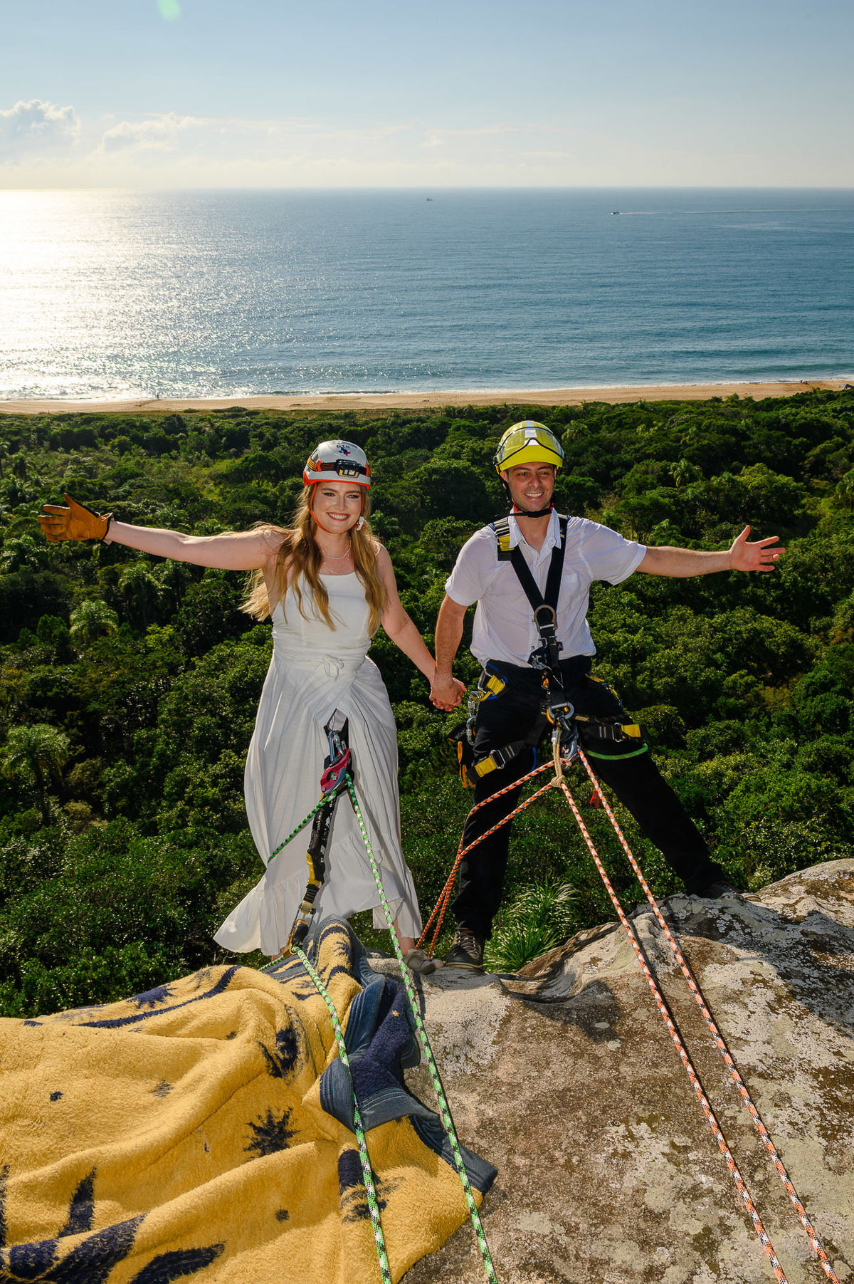 Casal de mãos dadas no topo da rocha, preparados para o rappel, com vista ampla do mar e da mata ao fundo