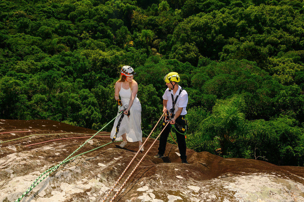 Casal descendo a rocha juntos no rappel, com o mar ao fundo e vegetação costeira ao redor