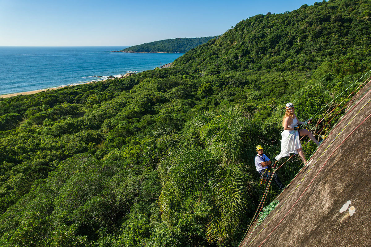 Vista ampla da rocha com o casal praticando rappel durante o ensaio pré-wedding, com o mar e a vegetação costeira ao fundo