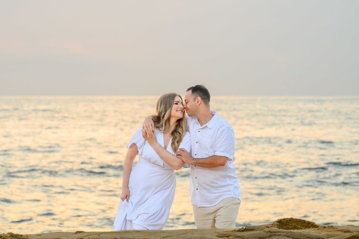 Casal caminhando próximo ao mar durante o amanhecer, com luz suave e reflexo dourado na água, ensaio pré-wedding na praia em Balneário Camboriú, SC
