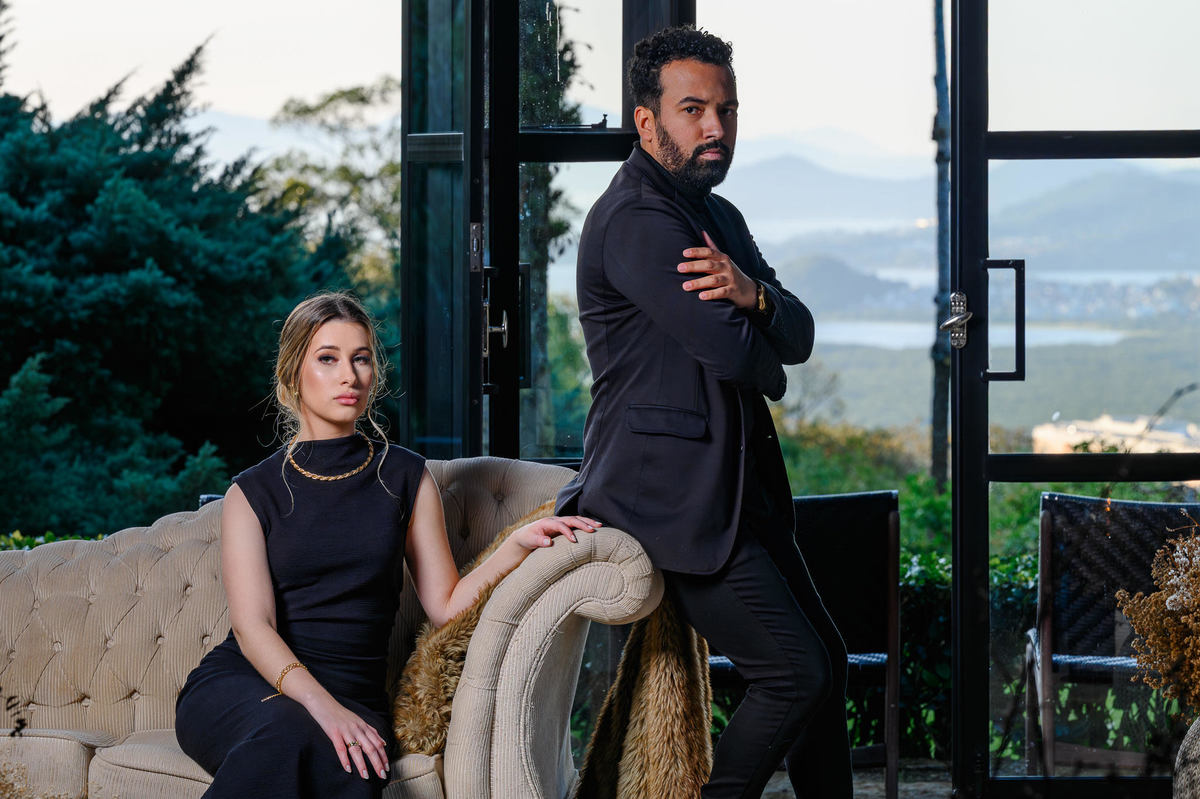 Casal em ensaio pré-wedding na Casa do Maciço em Florianópolis, posando em sofá com vista panorâmica ao fundo