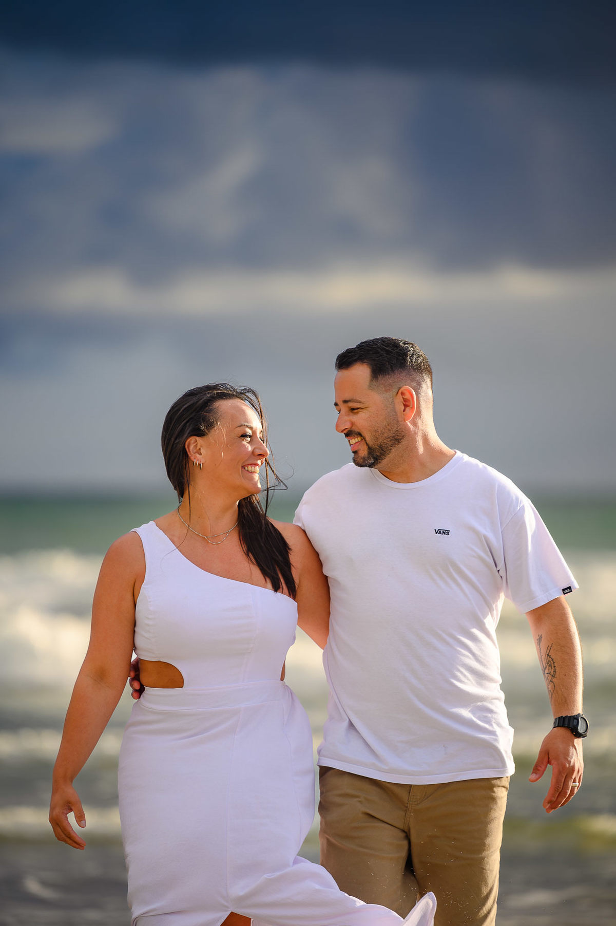 Ensaio de casal caminhando na praia de Bombinhas SC com céu dramático ao fundo, fotografia no litoral de Santa Catarina
