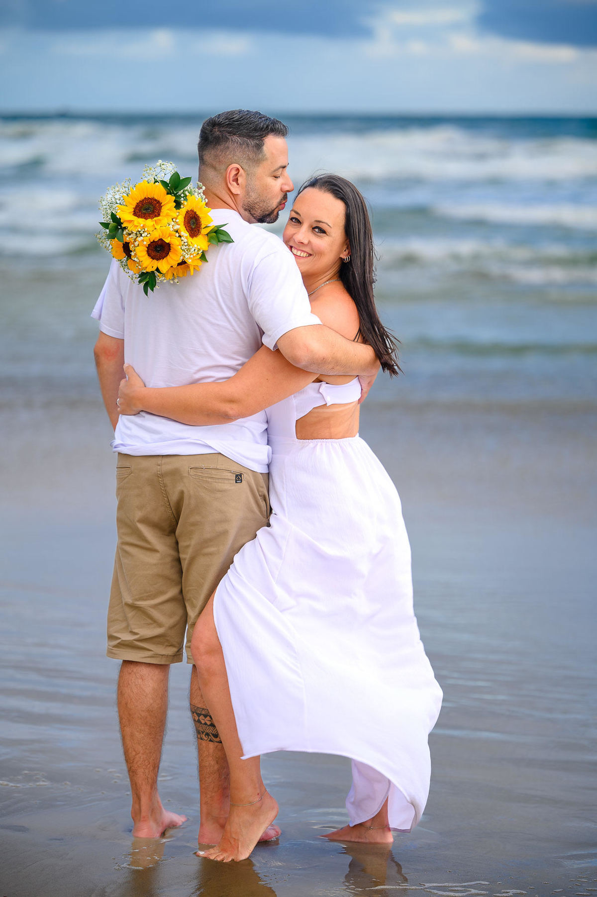 Casal abraçado na beira do mar em Bombinhas SC com buquê de girassóis, ensaio fotográfico de casal na praia