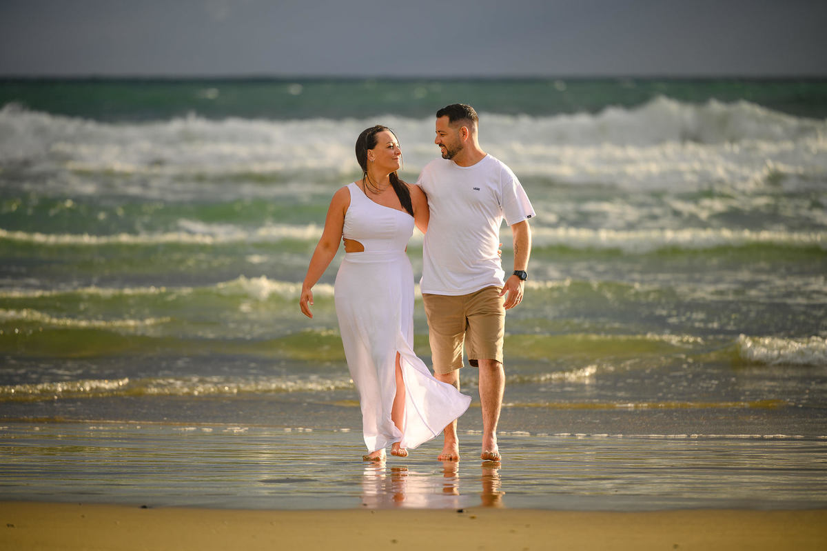 Casal caminhando na praia de Bombinhas SC com ondas do mar ao fundo, ensaio fotográfico de casal no litoral