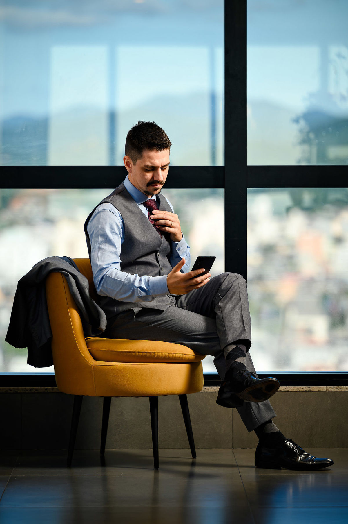 Retrato corporativo de advogado sentado em poltrona em ambiente moderno com vista para Balneário Camboriú, usando celular, fotografia profissional de posicionamento de imagem.