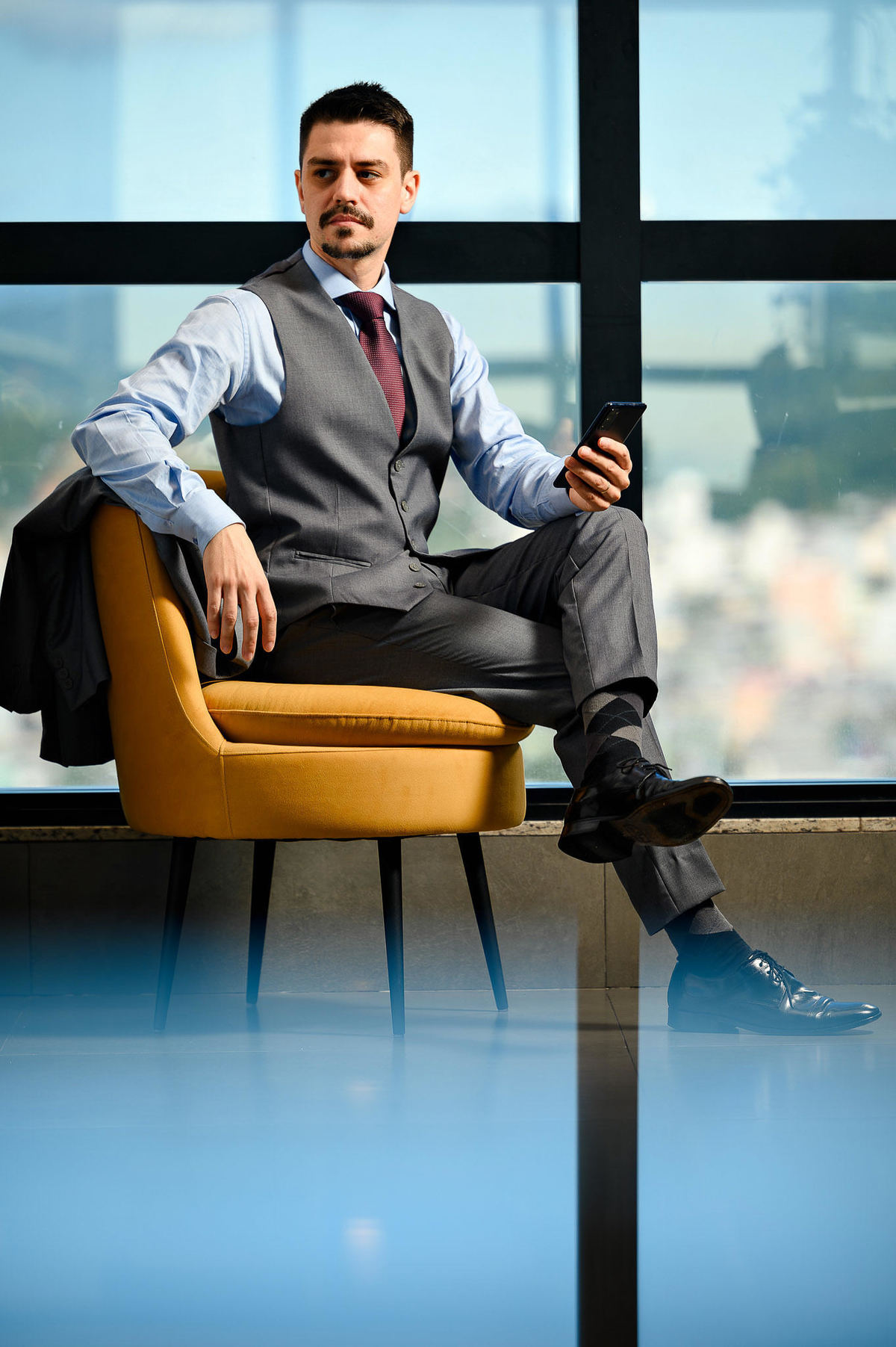 Retrato corporativo de advogado sentado em poltrona em ambiente moderno com vista para Balneário Camboriú, usando celular, fotografia profissional de posicionamento de imagem.