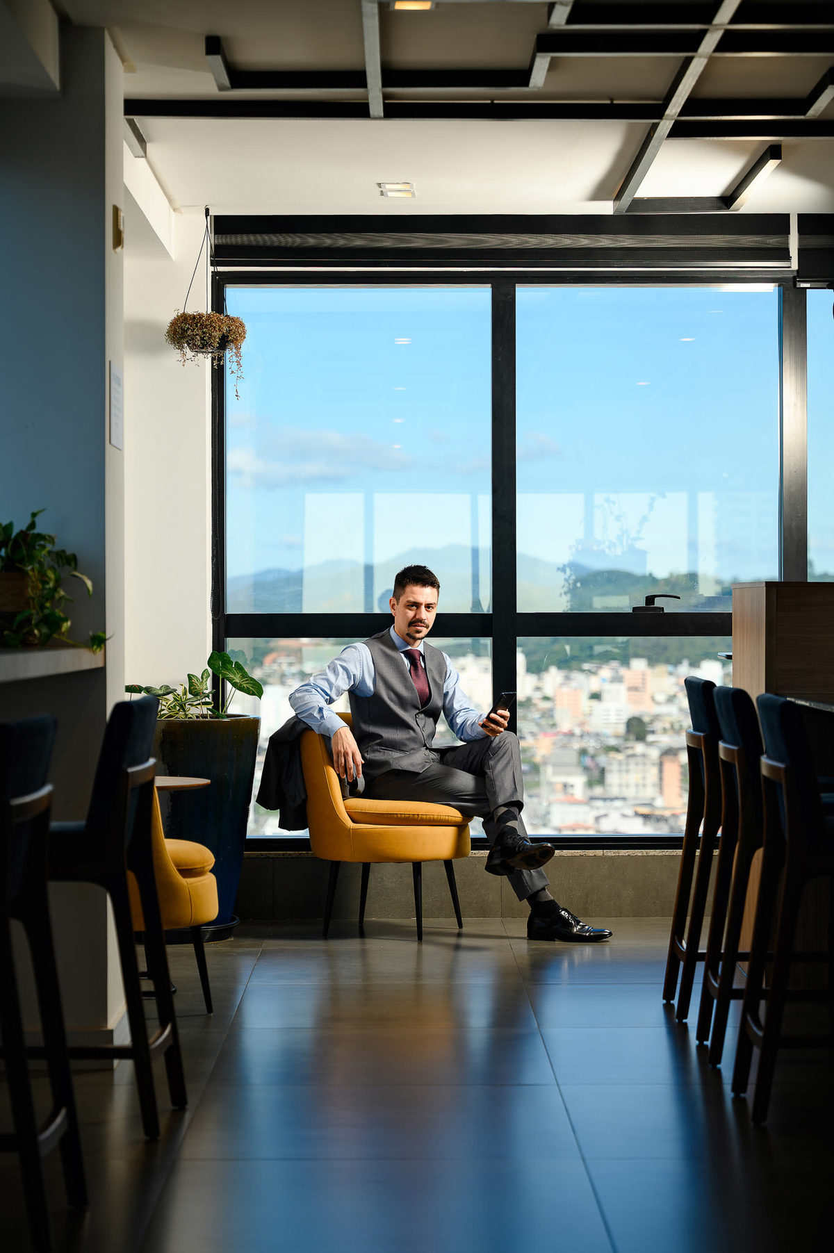 Retrato corporativo de advogado sentado em poltrona em ambiente moderno com vista para Balneário Camboriú, usando celular, fotografia profissional de posicionamento de imagem.