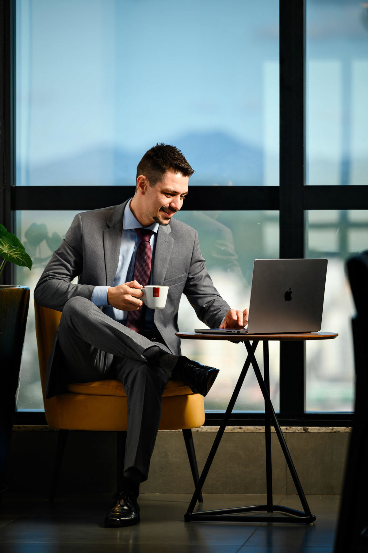 Retrato corporativo profissional de advogado sorrindo com xícara de café ao lado de notebook em ambiente corporativo com vista urbana em Balneário Camboriú.