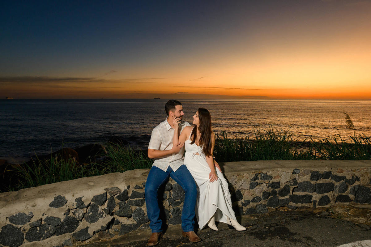 Casal sentado em mureta de pedra com vista para a Praia de Taquarinhas, em Balneário Camboriú, durante ensaio pré-wedding ao amanhecer