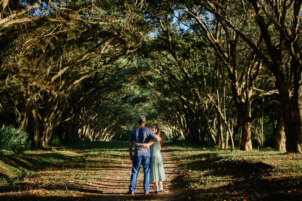 Casal caminhando de mãos dadas por estrada arborizada no bosque da Praia de Taquarinhas, em Balneário Camboriú, no ensaio pré-wedding