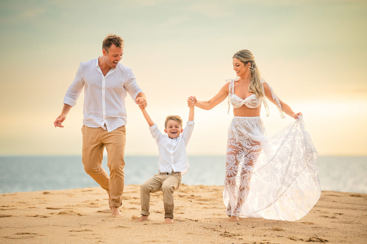 Família em ensaio pré-wedding ao amanhecer na praia em Itapema SC, com casal e filho caminhando na areia com luz suave da manhã.