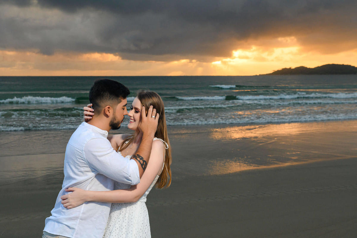 Casal se abraçando de frente para o mar ao amanhecer na praia de Bombas em Bombinhas, SC, com reflexo da luz da manhã na areia molhada
