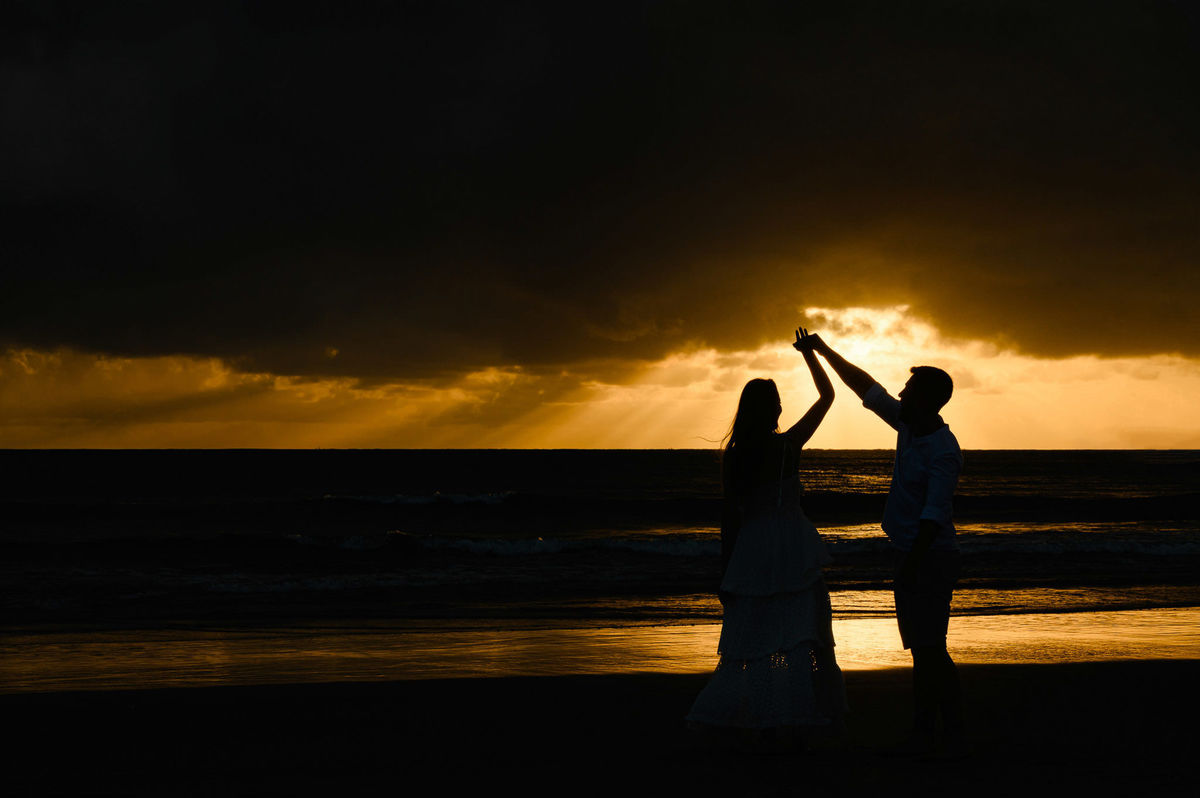 Casal em silhueta dançando de mãos dadas ao amanhecer na praia de Bombas em Bombinhas, com o sol surgindo entre as nuvens