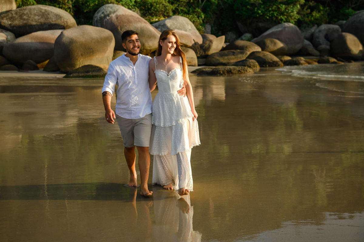 Casal caminhando entre pedras na praia de Bombas em Bombinhas ao amanhecer, com luz suave iluminando o cenário natural