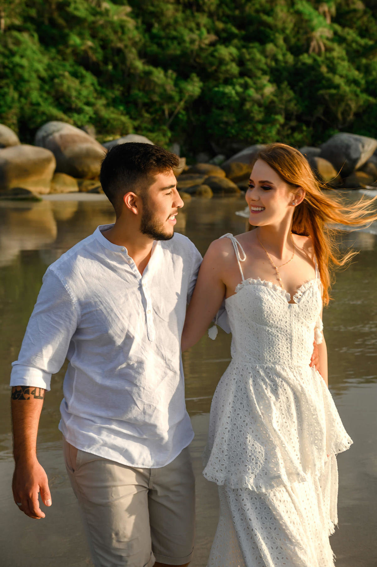 Casal caminhando entre pedras na praia de Bombas em Bombinhas ao amanhecer, com luz suave iluminando o cenário natural