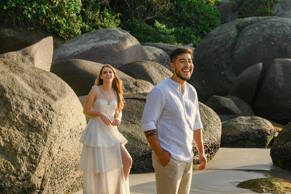 Casal andando entre as rochas da praia de Bombas em Bombinhas durante ensaio pré-wedding ao amanhecer, com mar calmo ao fundo